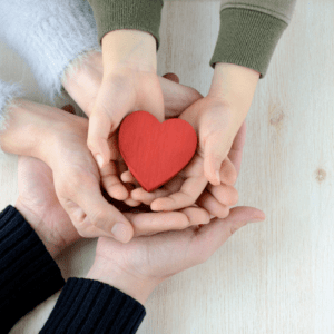 Three hands holding a wooden red heart
